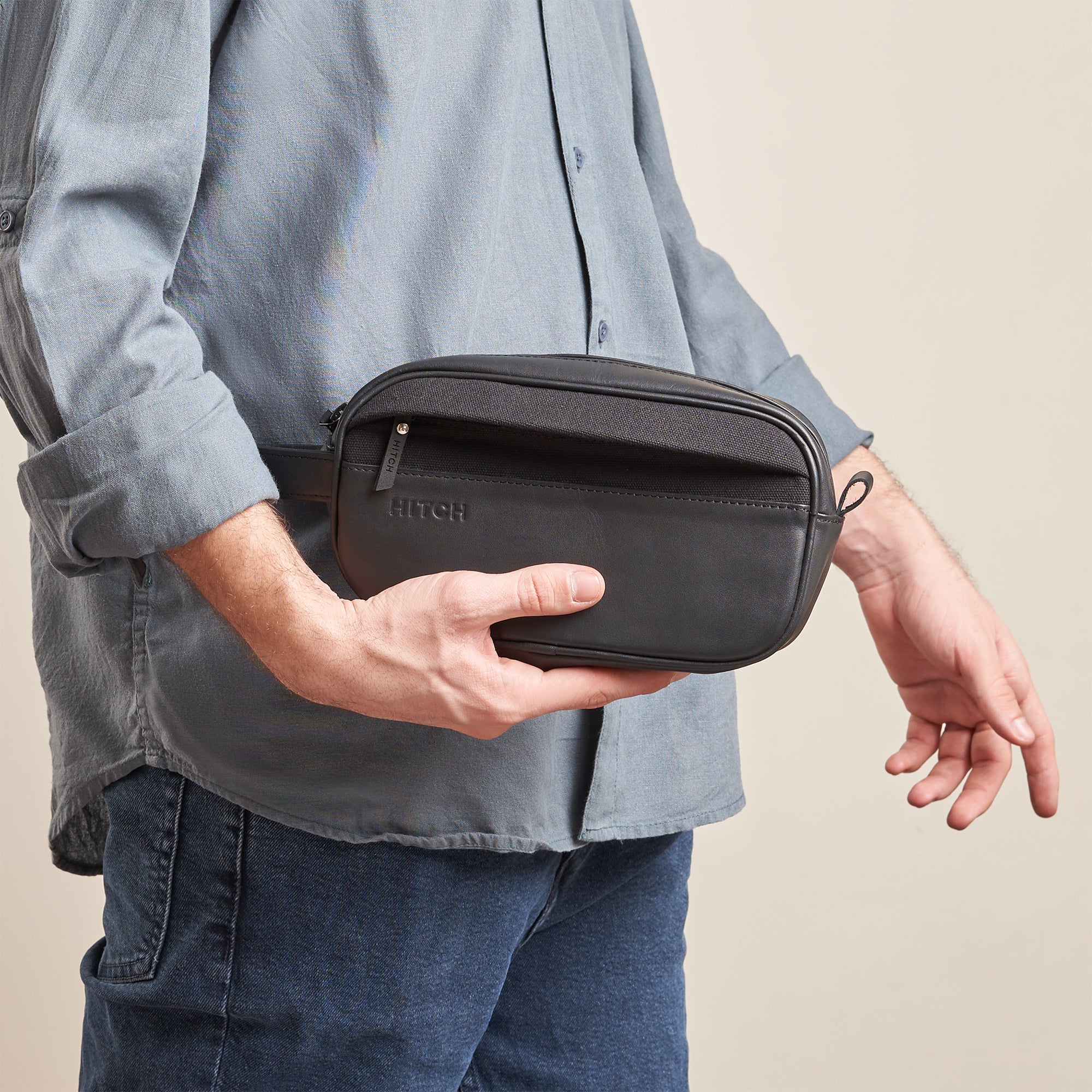 Man holding black leather dopp kit from HITCH, casual style, on a beige background.